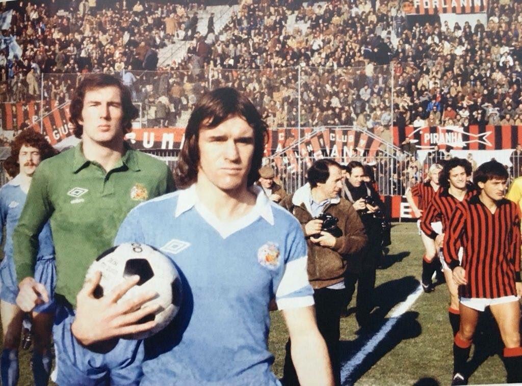 San Siro, 23 November 1978: Manchester City captain Dave Watson leads his side out — Joe Corrigan in green behind him, Milan's stripes following — before the UEFA Cup third-round first leg, played in afternoon light after fog had forced a postponement. It ended 2-2, Milan clawing back a two-goal deficit in front of terraces draped with Milan Club banners; the season of the star, seen from the Curva.