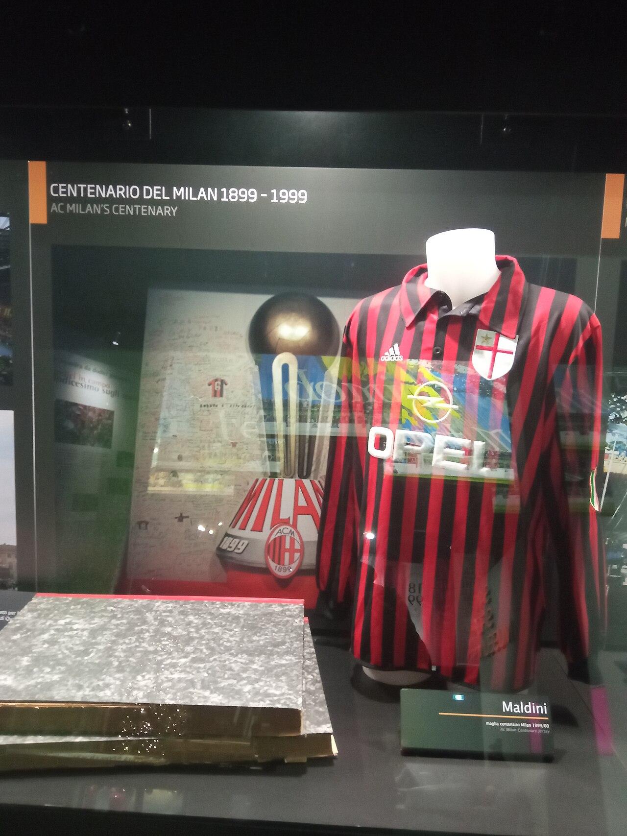 The San Siro museum's display case headed CENTENARIO DEL MILAN 1899-1999: Paolo Maldini's shirt, labelled by the curators "maglia centenario Milan 1999/00", hung beside the signed centenary board. Note the breast — no club badge, but the white shield and red cross of the city of Milan itself, worn for the hundredth year. The club had turned a hundred on 16 December 1999, six months after the captain lifted the sixteenth scudetto.